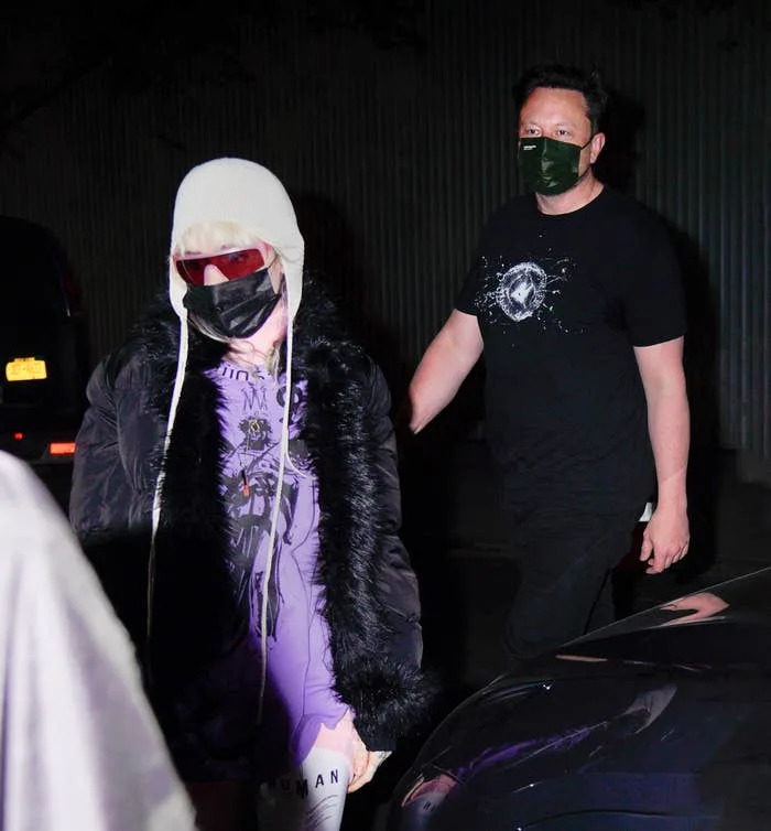 Grimes and Elon Musk wearing casual, dark outerwear and face masks walk along a dimly lit street at night. One has a distinctive hat and sunglasses