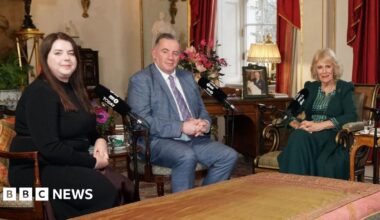 Amy Hunt, a woman with brown hair wearing a black long sleeved dress, and John Hunt wearing a grey checked suit sit with Queen Camilla who wears a dark green outfit.  Baroness Theresa May and Emma Barnett  were also sat around a table in Clarence House but out of shot.
