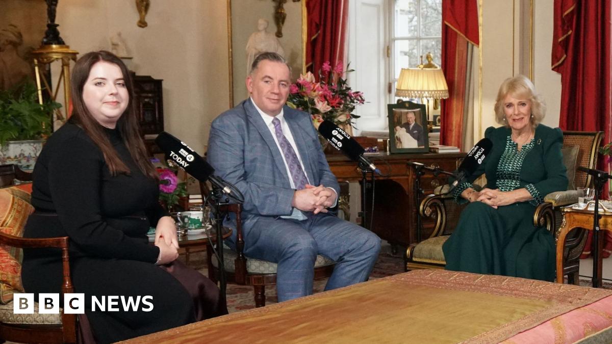 Amy Hunt, a woman with brown hair wearing a black long sleeved dress, and John Hunt wearing a grey checked suit sit with Queen Camilla who wears a dark green outfit.  Baroness Theresa May and Emma Barnett  were also sat around a table in Clarence House but out of shot.