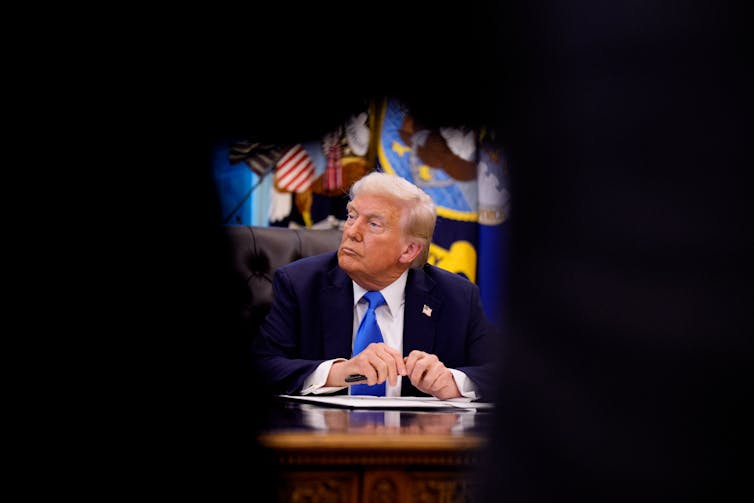 A man in a dark suit and blue tie sits at a desk.