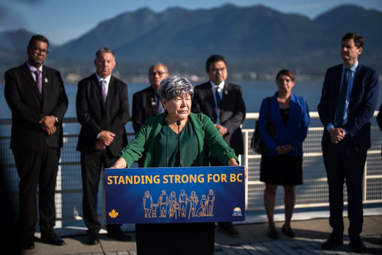 an older woman with greying hair speaks at a podium, people in suits stand behind her. mountains are in the background