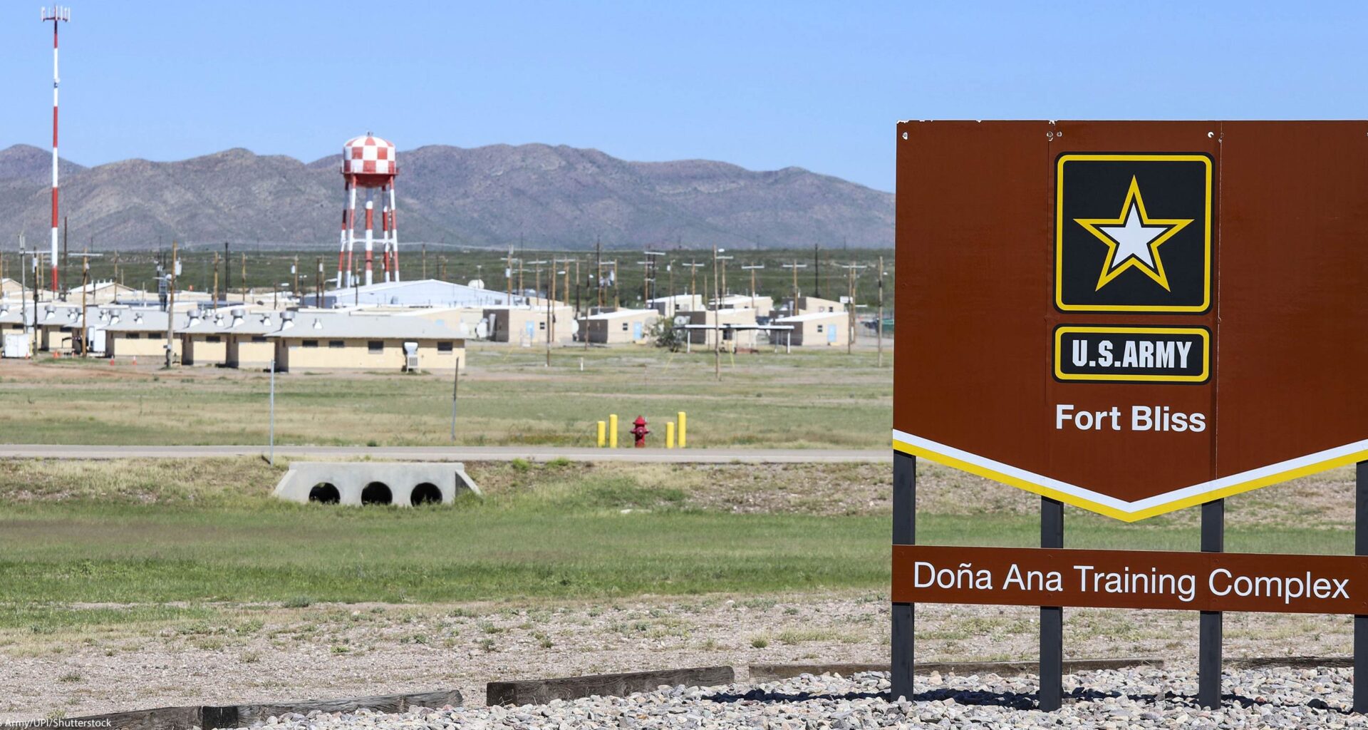 A large brown U.S. Army sign reading “Fort Bliss – Doña Ana Training Complex” stands in the foreground of a wide, open landscape. Behind it are low beige military buildings, utility poles, and a tall red-and-white checkered water tower, with mountains rising in the distance under a clear blue sky.