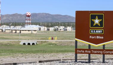 A large brown U.S. Army sign reading “Fort Bliss – Doña Ana Training Complex” stands in the foreground of a wide, open landscape. Behind it are low beige military buildings, utility poles, and a tall red-and-white checkered water tower, with mountains rising in the distance under a clear blue sky.