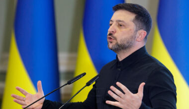 Volodymyr Zelensky speaks at a podium, gesturing with open hands, with Ukrainian flags in the background.