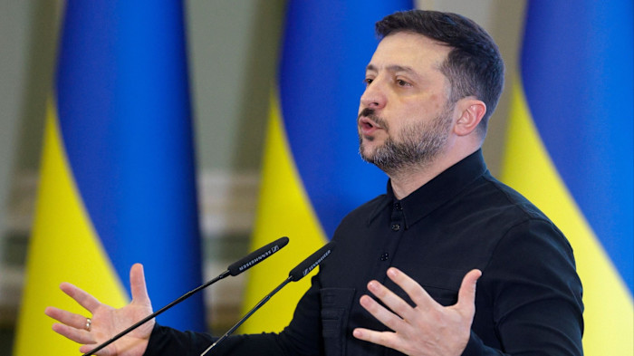 Volodymyr Zelensky speaks at a podium, gesturing with open hands, with Ukrainian flags in the background.