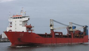 The Russian cargo ship Adler sails on the water, showing a red hull, white superstructure, and onboard cranes.