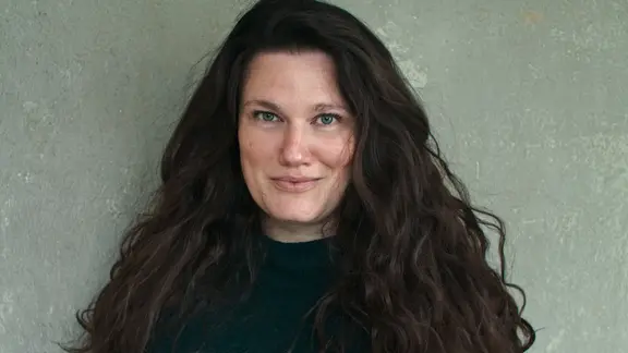 Eine Frau mit langen dunklen Haaren schaut etwas schüchtern in eine Kamera.