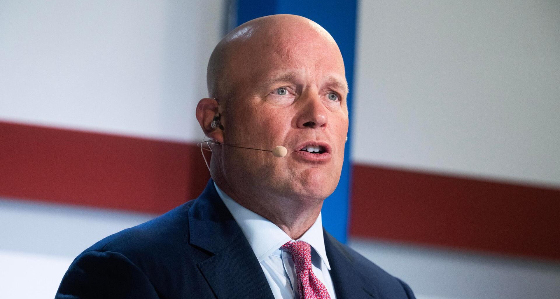 Der glatzköpfige Whitaker mit dunklem Jackett und roter Krawatte sagt etwas. Dahinter eine weiße Wand mit einem roten Quer- und einem blauen Längstreifen, die sich kreuzen.