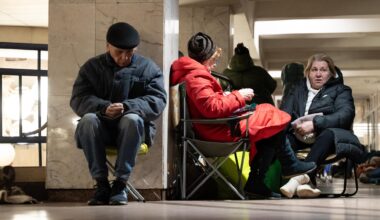 Ein älterer Mann sitzt in Winterkleidung auf einem Klappstuhl an eine Wand gelehnt und scheint zu schlafen. Neben ihm zwei Frauen auf Klappstühlen.