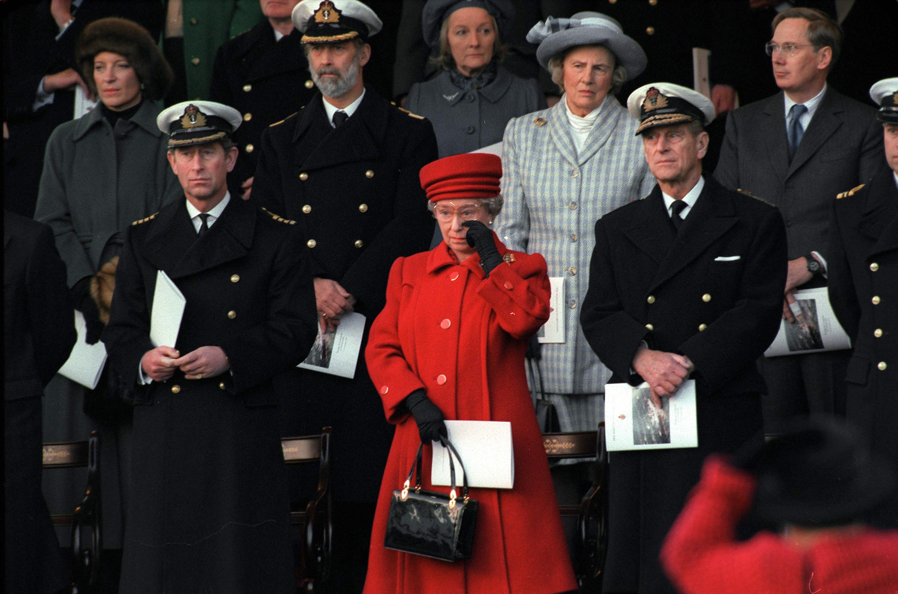 Queen Elizabeth wearing a red coat crying standing next to Prince Charles and Prince Philip