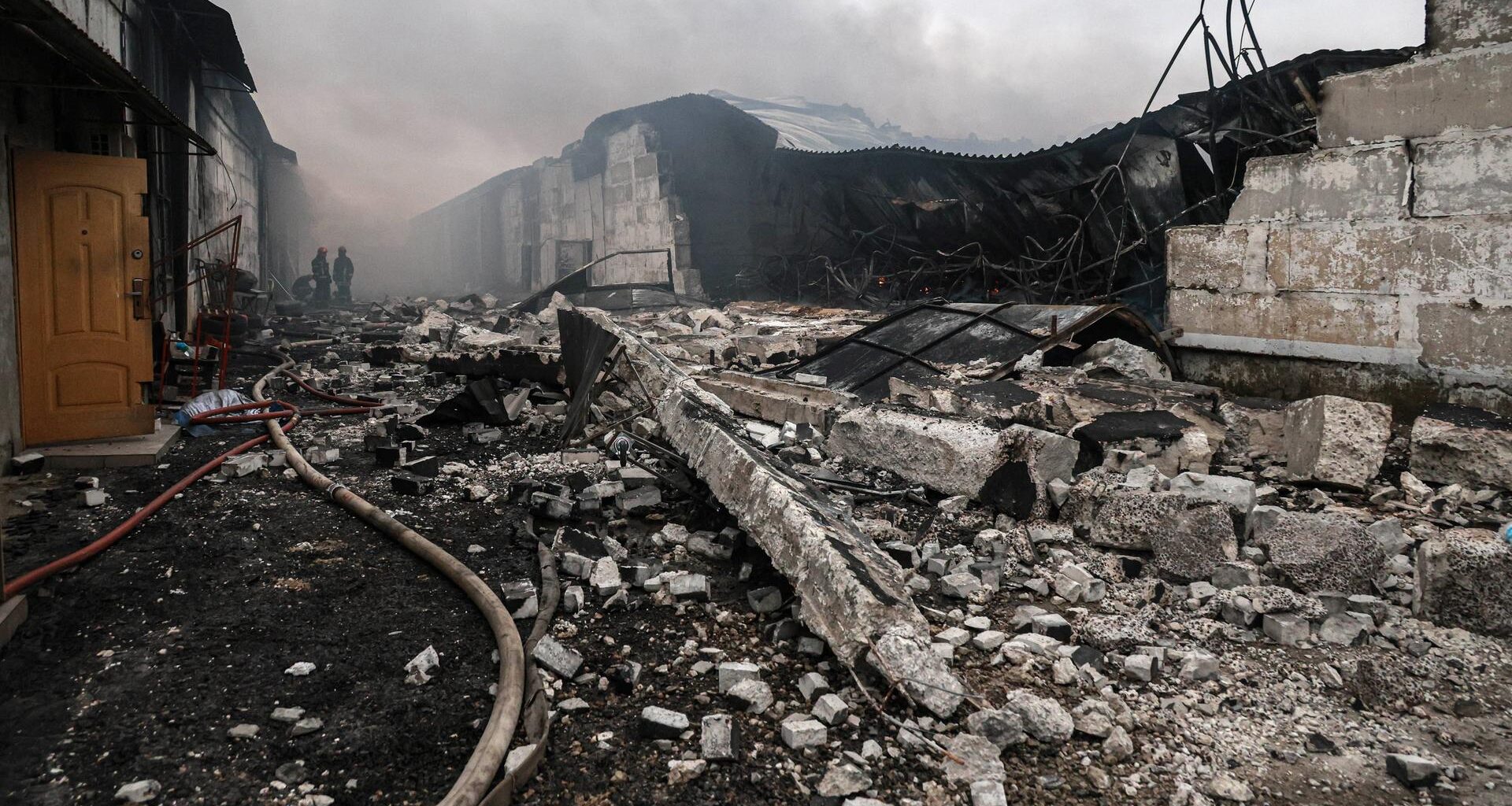 Das Bild zeigt ein Trümmerfeld aus Beton. Ein Löschschlauch führt zu zwei Feuerwehrmännern im Bildhintergrund, darüber Rauch.