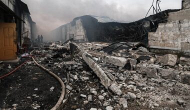Das Bild zeigt ein Trümmerfeld aus Beton. Ein Löschschlauch führt zu zwei Feuerwehrmännern im Bildhintergrund, darüber Rauch.