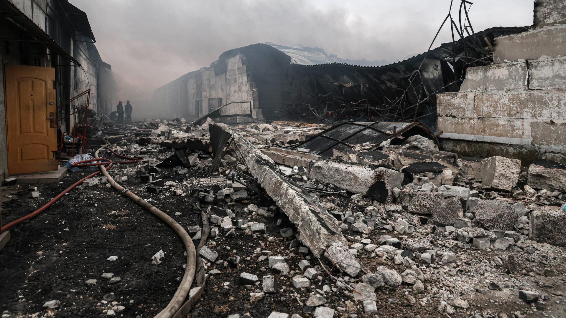 Das Bild zeigt ein Trümmerfeld aus Beton. Ein Löschschlauch führt zu zwei Feuerwehrmännern im Bildhintergrund, darüber Rauch. 