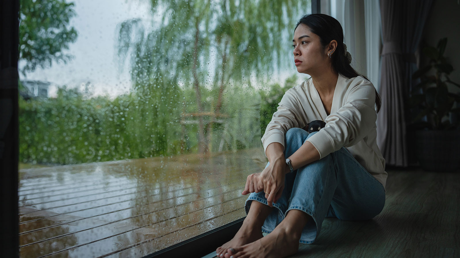 Woman with worried expression looking out window at a rainy outdoor scene