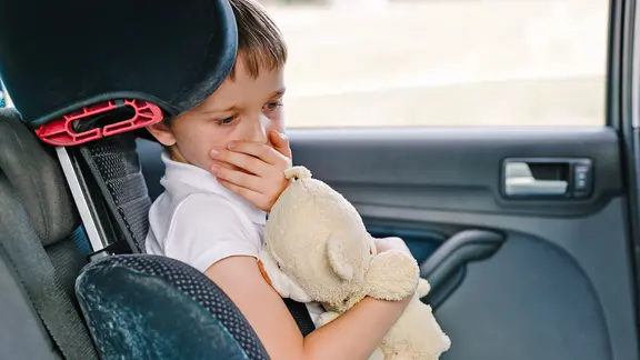 Kind sitzt mit Teddy im Arm im Auto und hält sich die Hand vor den Mund.