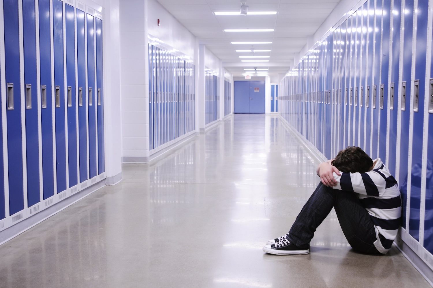 boy sitting with head in hands in school corridor amid blue lockers
