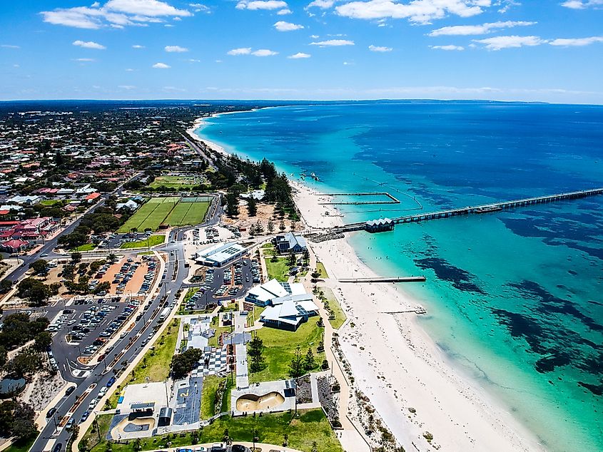 An aerial view of Busselton Jetty in Western Australia reveals the iconic wooden pier extending over the turquoise waters, with the shoreline and town of Busselton visible in the background.