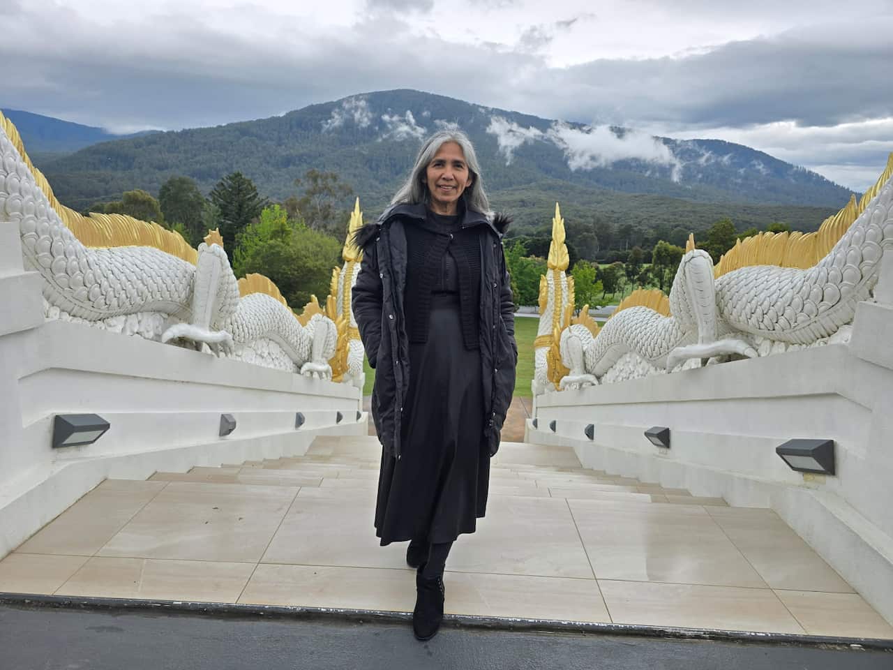 A woman with long grey hair, wearing a long black coat and dress, stands smiling on wide white stairs flanked by decorative white and gold dragon statues, with a large, misty, tree-covered mountain in the background under a cloudy sky.