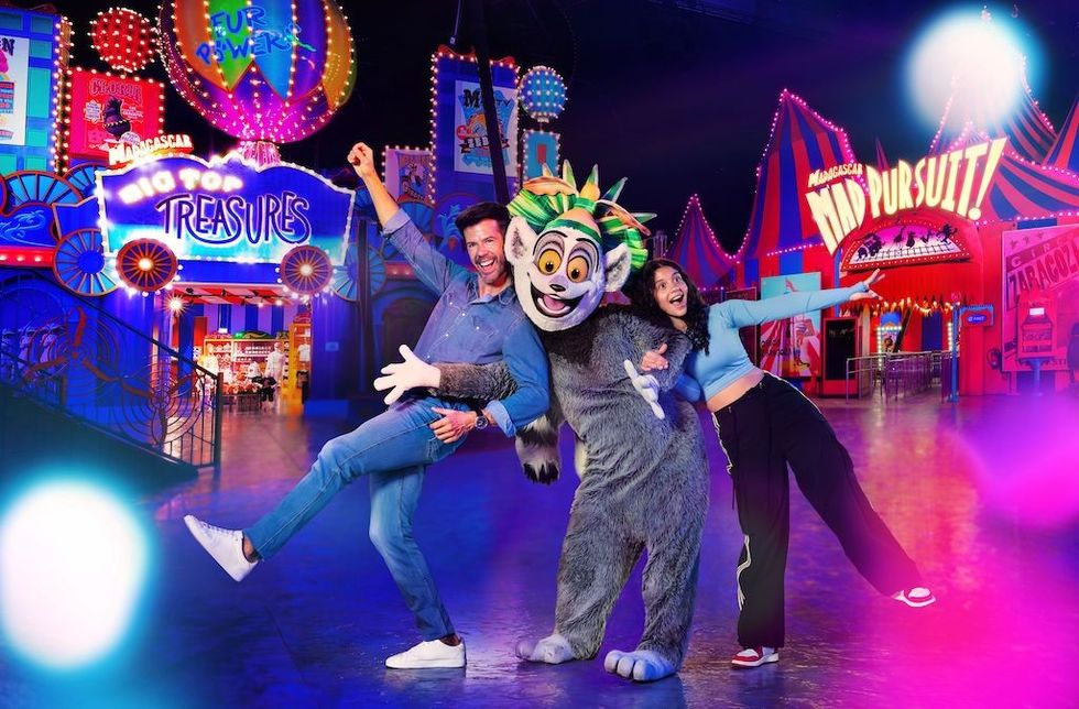 Two people pose with a lemur mascot in a vibrant, colorful amusement park setting.