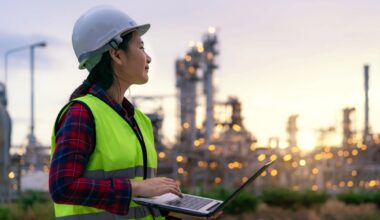 A worker in a hard hat and green reflecting vest looks at an industrial power plant while holding a laptop.