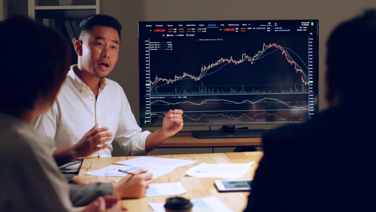 A person talking to two others with a price chart in the background.