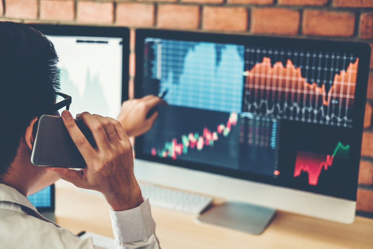 A person talks on a phone while pointing at a computer screen that displays stock price charts.