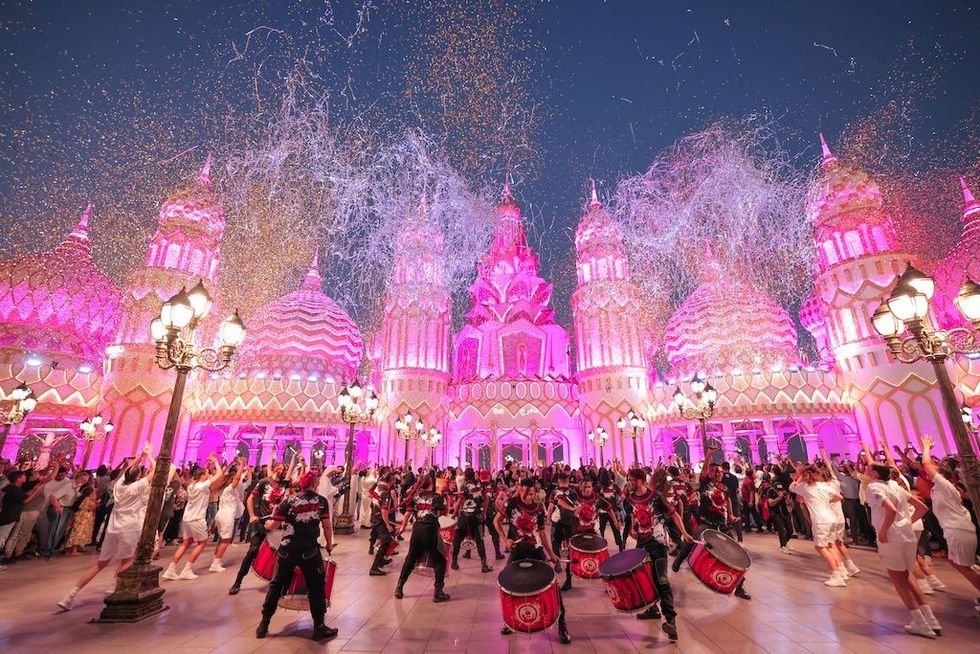 Vibrant celebration with performers and fireworks in front of a pink-lit ornate building.