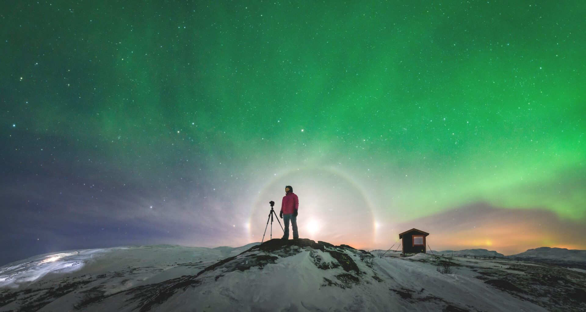 En person står framför en ljusbåge på en snötäckt klippa under en norrskensgrön himmel.