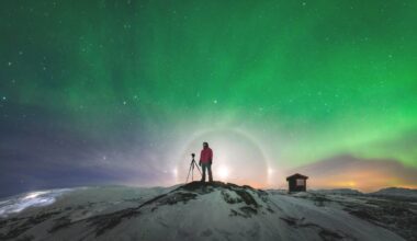 En person står framför en ljusbåge på en snötäckt klippa under en norrskensgrön himmel.