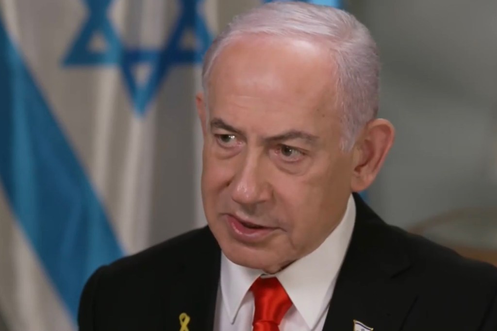 Benjamin Netanyahu in a black suit, red tie, and a yellow ribbon, speaking in front of an Israeli flag.
