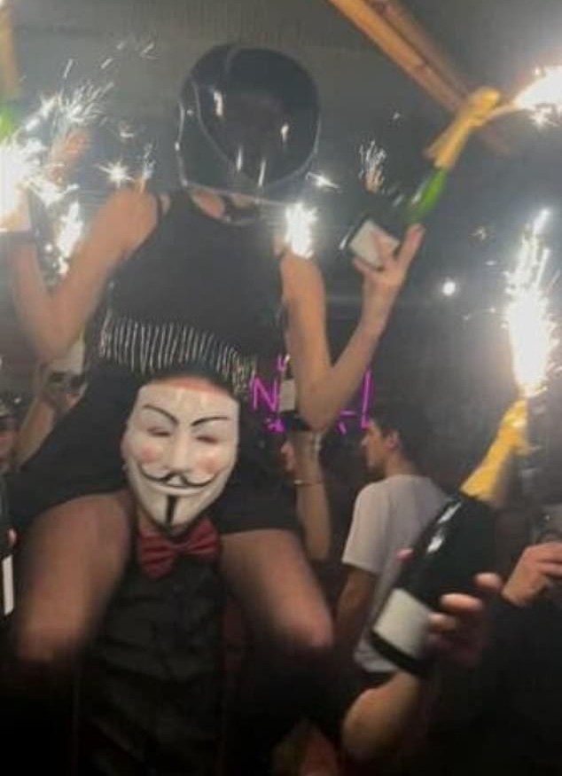 A person on another's shoulders, holding two bottles of sparkling wine with roman candle fireworks, at a bar in Crans Montana.