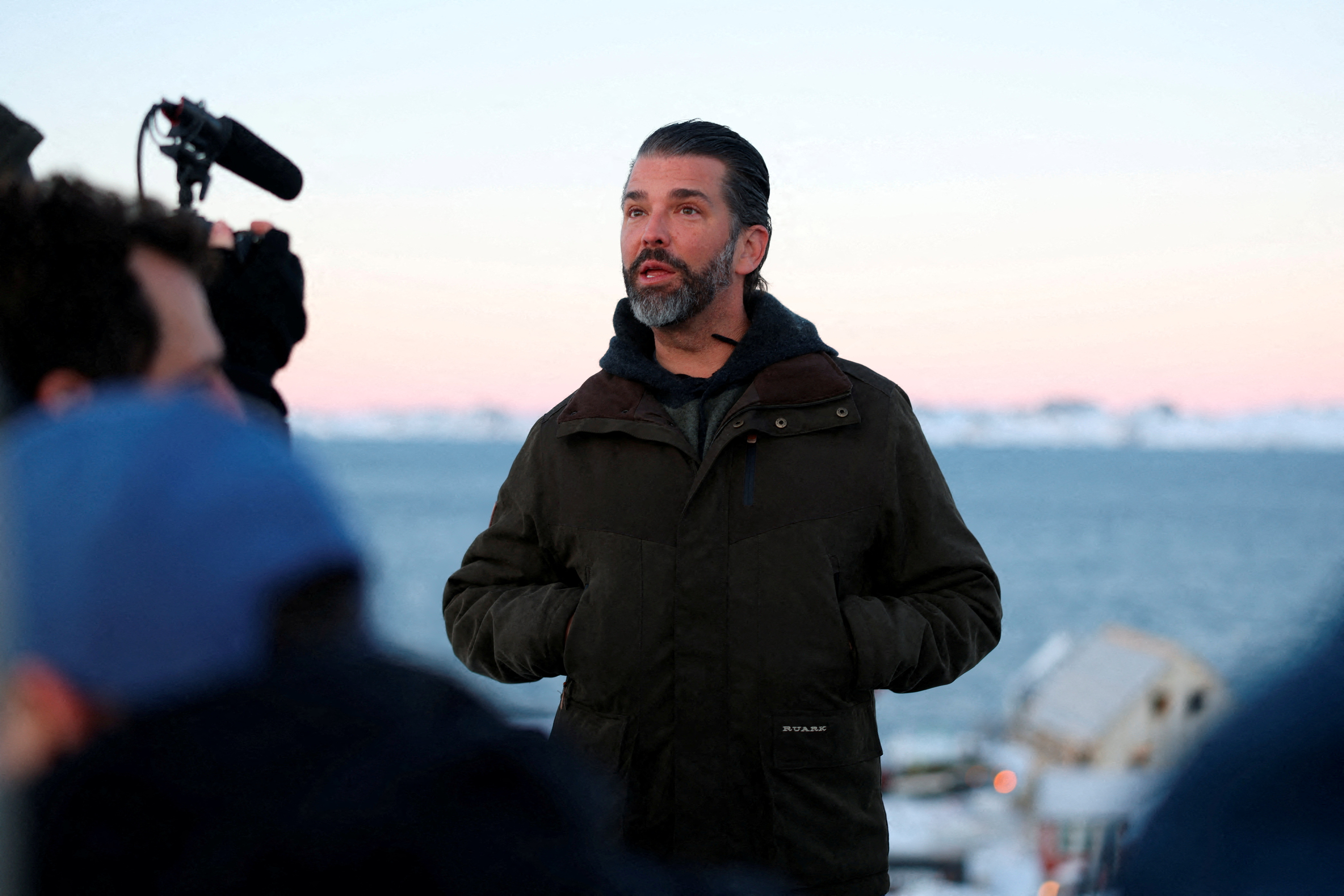 Donald Trump Jr. in Nuuk, Greenland, wearing a dark jacket, looking upwards, with a microphone visible in the foreground.