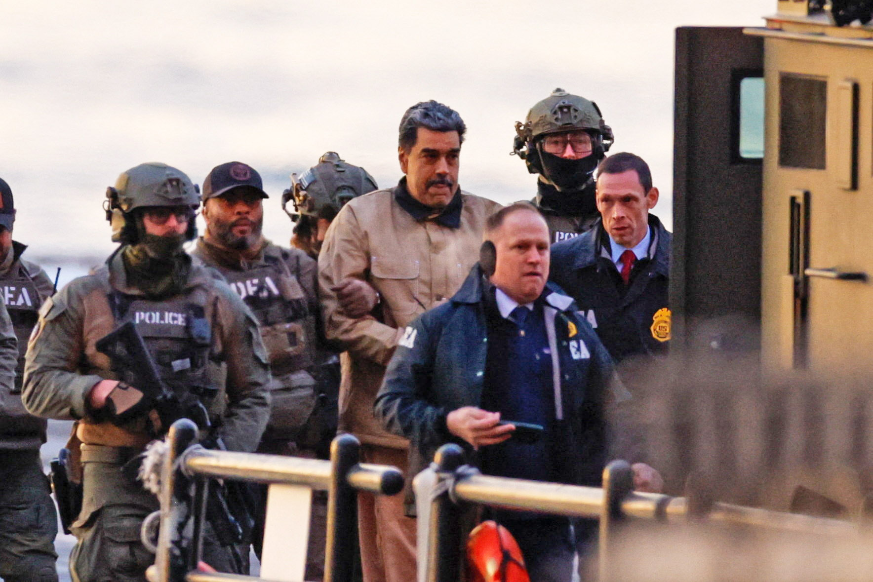 Nicolas Maduro, in a tan jacket, being escorted by law enforcement personnel in tactical gear and suits.