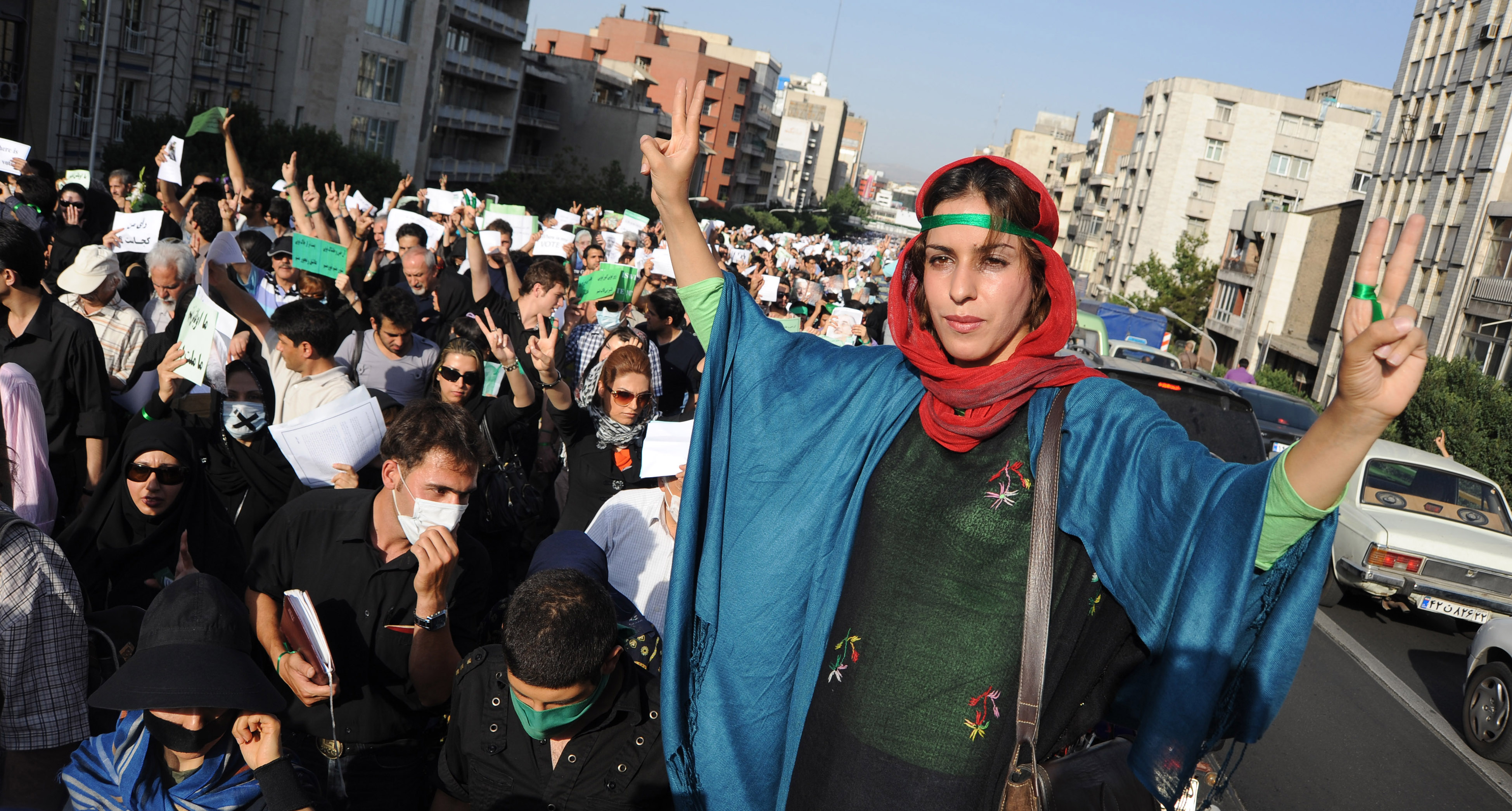 A female demonstrator with a red headscarf and blue shawl flashes a V-sign at a street protest.