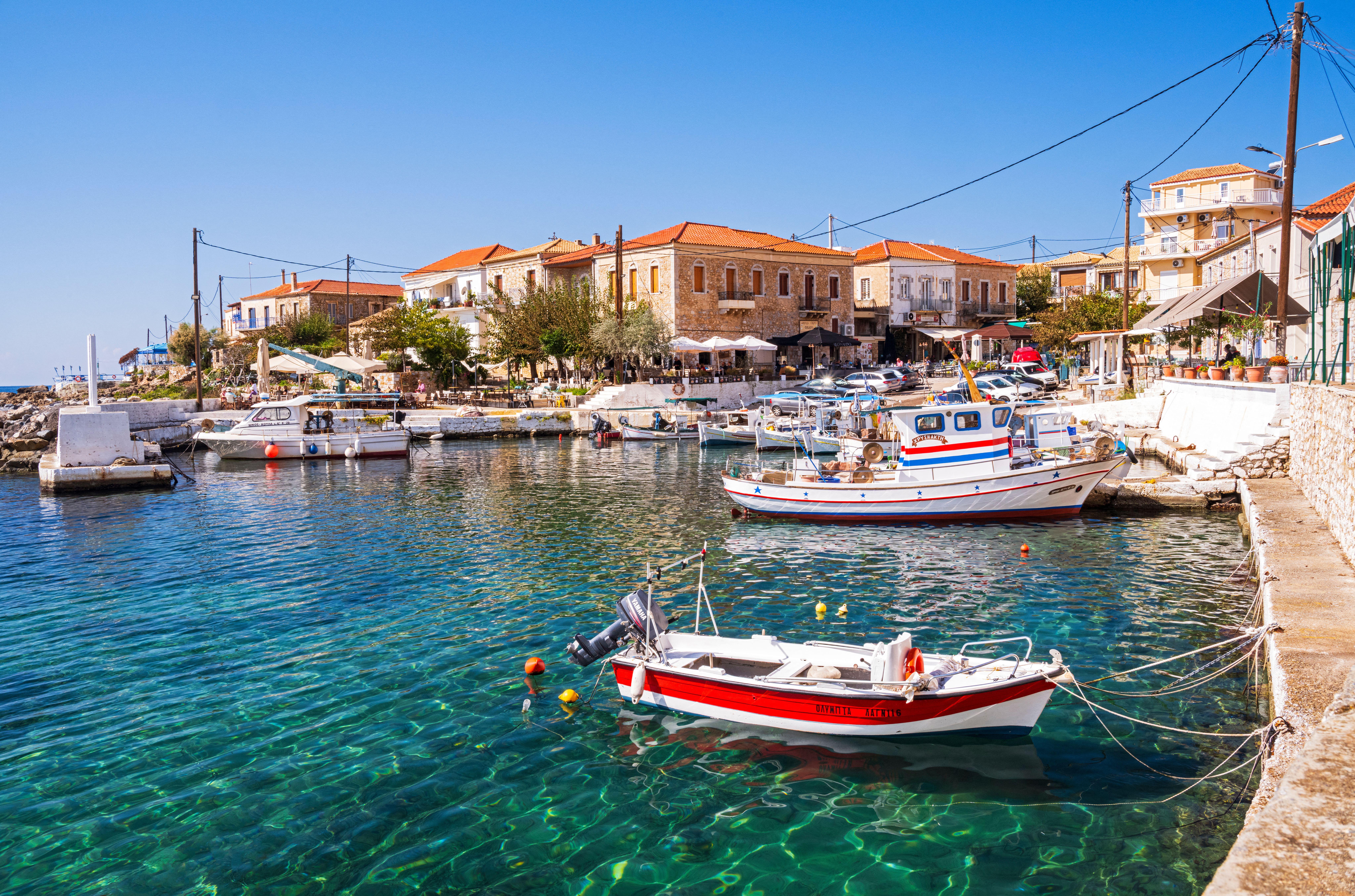 Fishing boats docked in the turquoise water of a harbor in Agios Nikolaos, Greece, with buildings and shops lining the shore.