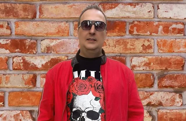 DJ AliPink wearing sunglasses, a red jacket, and a black t-shirt with a skull and roses design.