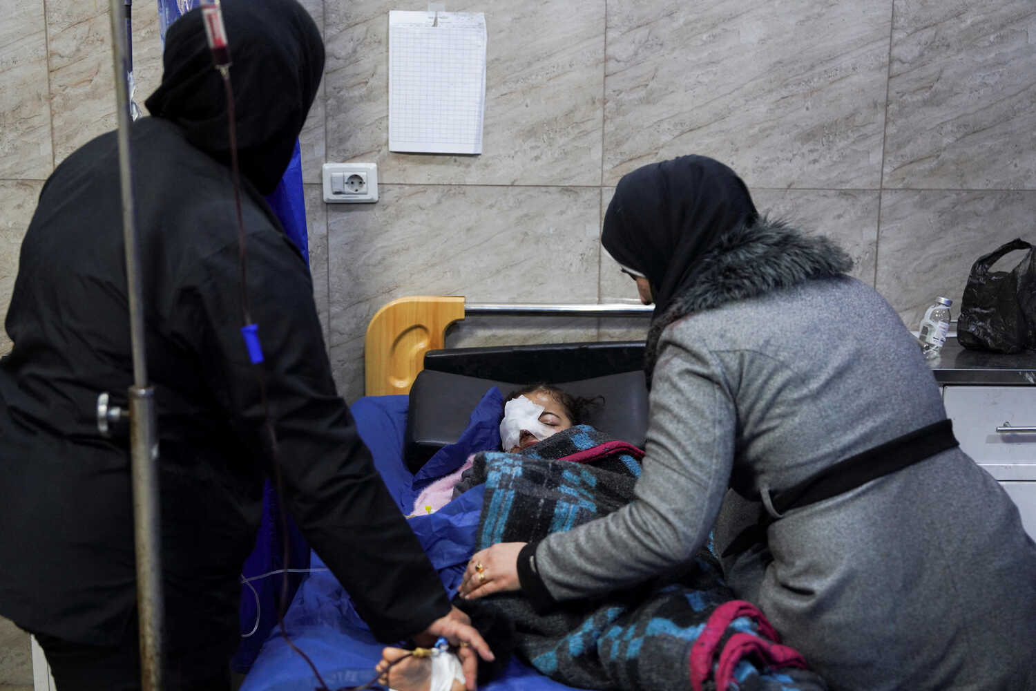 A child with a bandaged face rests in a hospital bed. Two people stand close by, one near an IV pole.