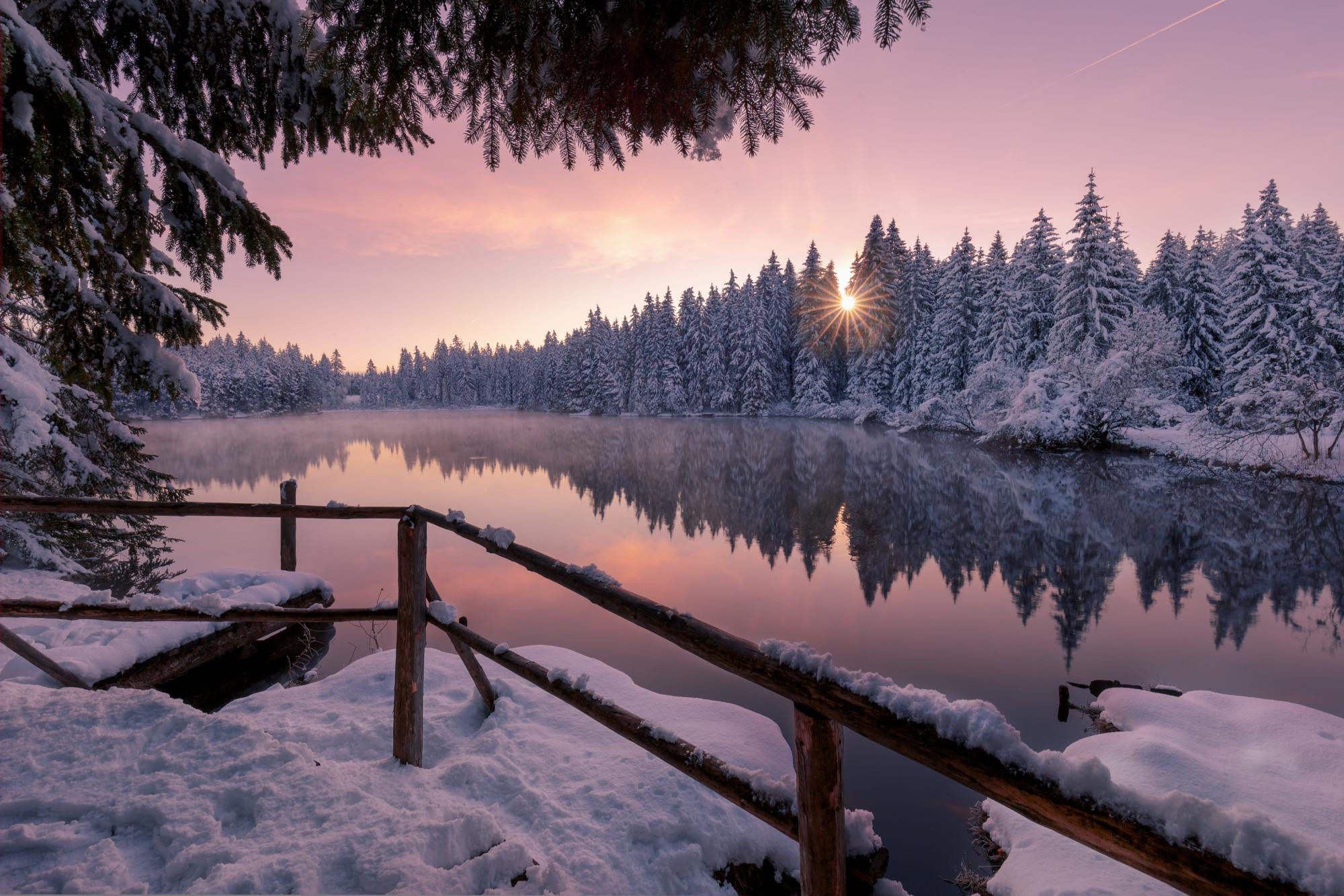 Lac enneigé entouré de sapins sous un ciel rose au coucher du soleil, reflet dans l’eau calme.