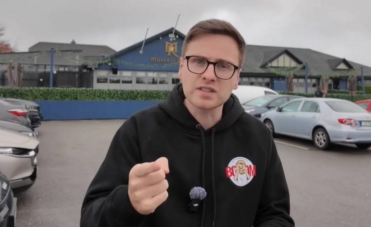 An individual dressed in a black hoodie, wearing glasses, and gesturing with their hand in an outdoor parking lot, with several cars parked in the background and a building with blue walls visible in the distance.