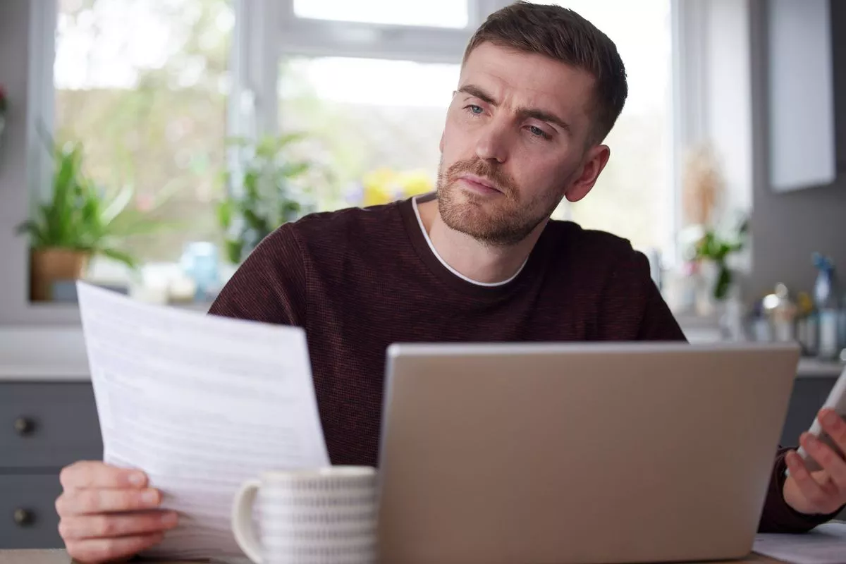 Man In Kitchen At Home Looking At Domestic Bills With Laptop And Calculator
