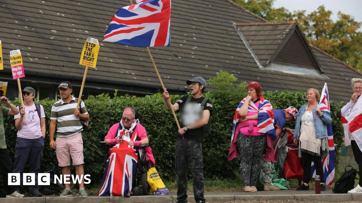 Protestors waving flags and counter protesters with signs saying 'Stop the far right' gather outside a hedge with a hotel behind it