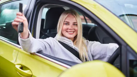 Getty Images A young woman smiles as she sits in a yellow car with her seatbelt on holding the keys out the window.