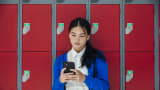 A medium close-up front view of a teenage girl who is sending text messages to a friend and checking her social media as she waits in the corridor of the high school she attends. 