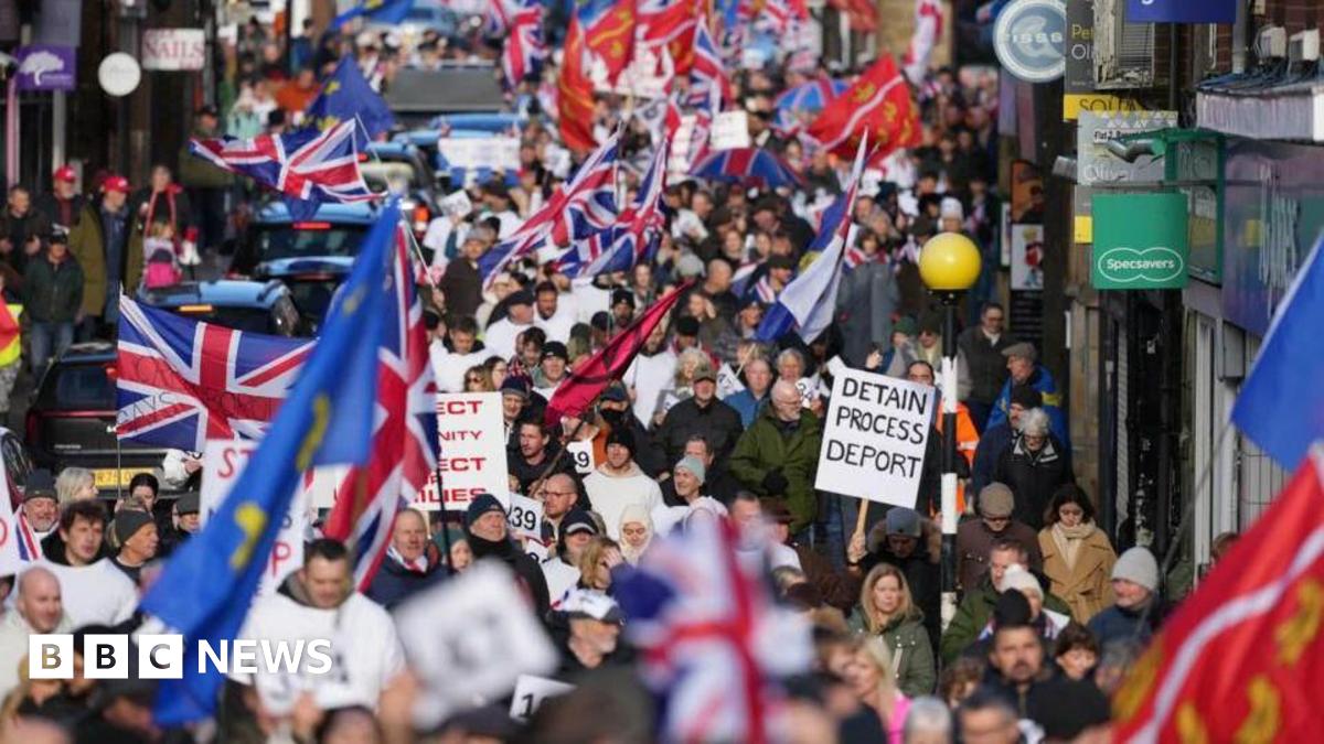 Crowds walk down the road in a protest waving Union flags. A banner says "detain, process, deport". There are shop signs to one side of the town centre road.