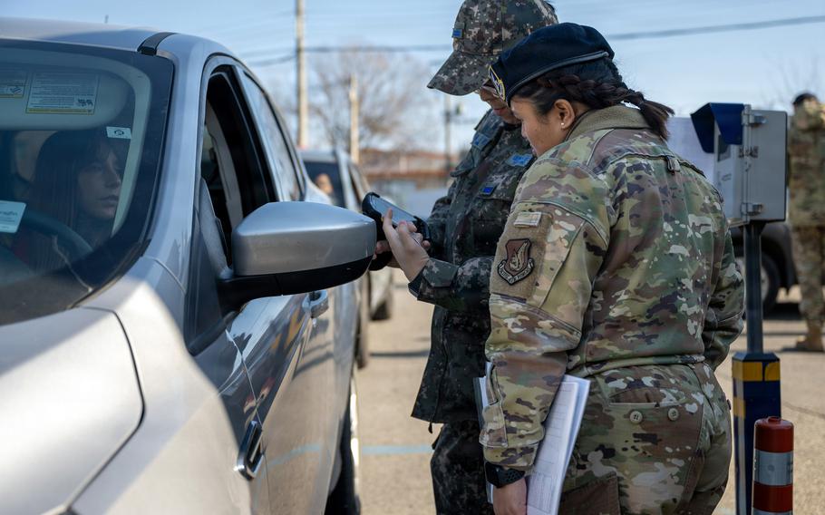 Two military personnel in camouflage uniforms conduct a vehicle inspection at a checkpoint, with one officer checking documents while speaking with the driver of a vehicle.