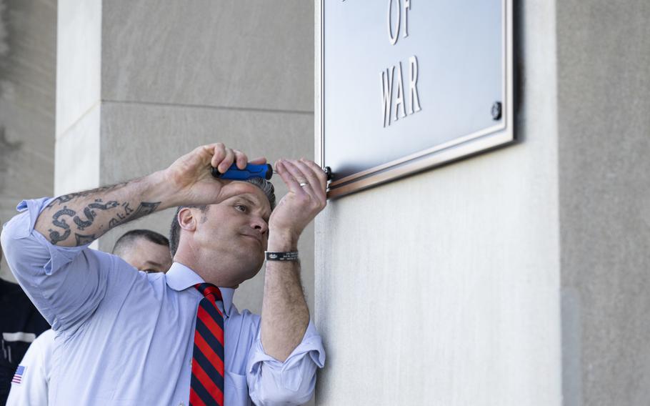Man in a light blue shirt and red-and-blue striped tie uses a screwdriver on a plaque mounted on a wall. 