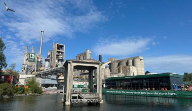 En stor sementfabrikk med siloer og industristrukturer i nærheten av et vann; på et grønt skilt står det: "The journey to net-zero concrete starts here." En måke flyr over bildet.