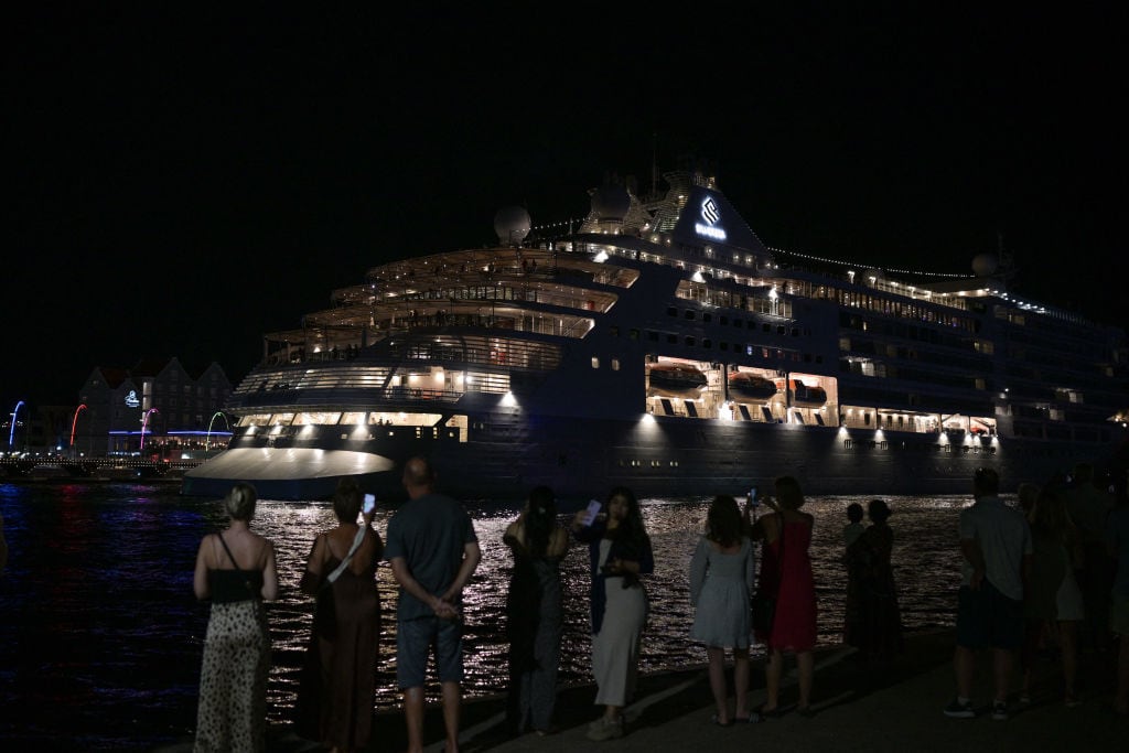 A group of people in vacation clothes stand in the dark watching a large cruise ship