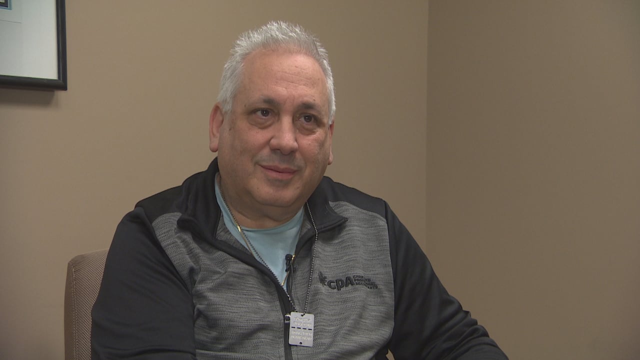 A man with white hair and wearing a zip-up grey jacket sits in a chair