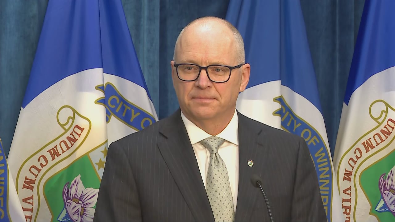 Bald man with dark eyeglasses wears a suit and stands infront of blue and white flags